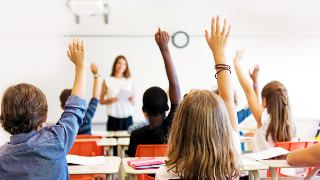 Lehrerin steht vor einer Schulklasse, die Kinder melden sich. Sie sind mit dem Rücken im Bild.